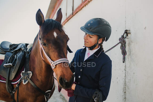 Vista lateral de joven adolescente mujer en casco de jinete y chaqueta acariciando caballo de pie juntos al aire libre - foto de stock
