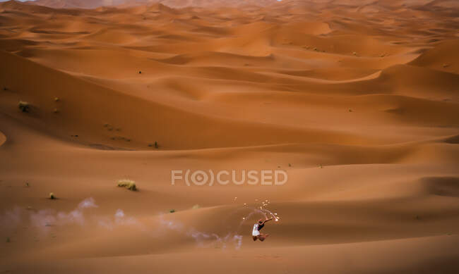 Vista aérea lejana de la alegre morena elegante sosteniendo el brazo levantado con fuegos artificiales ahumados en el desierto de Marruecos - foto de stock
