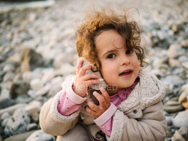 Retrato de linda niña escuchando concha con atención arrebatada mientras descansa en la playa pedregosa - foto de stock