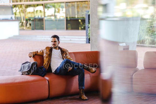Человек в модном наряде сидит на удобном красном кожаном диване в современном здании — стоковое фото