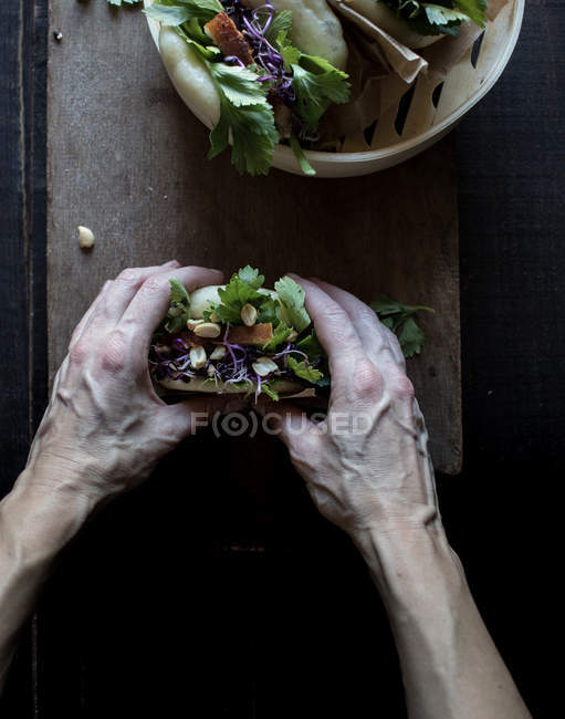 Von oben von den Händen, die leckeres Gua-Bao-Sandwich mit Speck, Erdnüssen und frischer Petersilie halten — Stockfoto