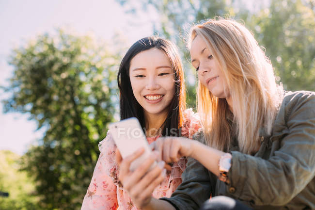Junge kaukasische Frau zeigt einem lächelnden asiatischen Freund ihr Smartphone, während sie an einem sonnigen Tag auf dem verschwommenen Hintergrund des Parks sitzt — Stockfoto