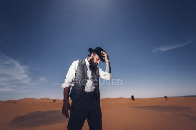 Homem barbudo em traje de cowboy olhando para baixo enquanto estava no deserto contra o céu azul — Fotografia de Stock