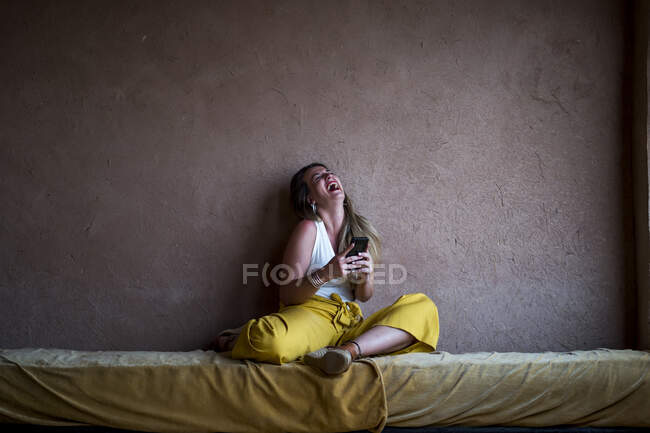 Happy blonde woman sitting on sofa on terrace in oriental style and using a mobile phone in Morocco — Stock Photo