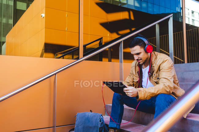 Schöner Mann mit Kopfhörern, der Musik hört und auf dem Tablet surft, während er auf der Treppe vor dem modernen Gebäude sitzt — Stockfoto