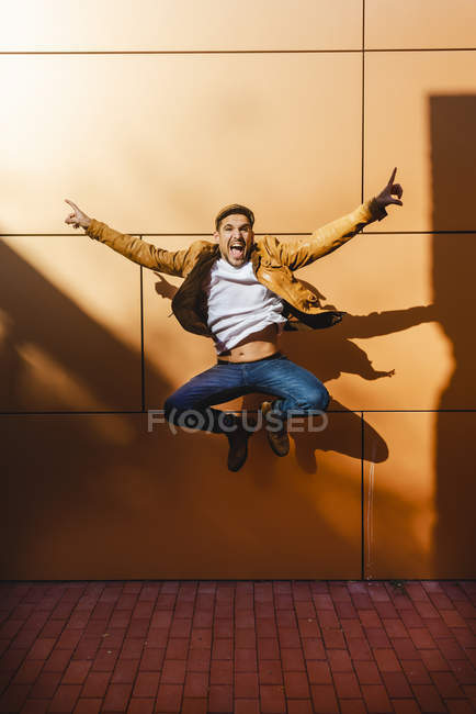 Joven positivo en traje elegante saltando cerca de la pared del edificio moderno en el día soleado - foto de stock