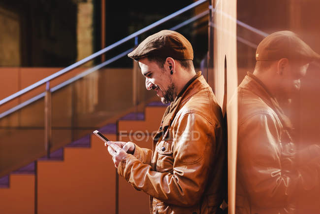 Hombre positivo en traje elegante usando el teléfono móvil mientras se apoya en la pared del edificio moderno en el día soleado - foto de stock