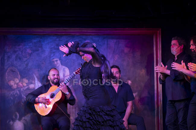 Mulher de traje preto dançando flamenco perto de músicos masculinos hispânicos durante a performance contra a pintura no palco escuro — Fotografia de Stock
