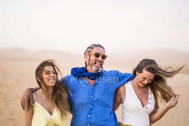 Старший бородатый мужчина смеется и обнимает веселых женщин во время прогулки по песчаной пустыне во время поездки в Марокко — стоковое фото