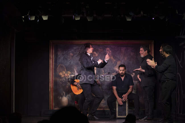 Человек в черном костюме танцует фламенко возле латиноамериканских музыкантов во время выступления против живописи на темной сцене — стоковое фото