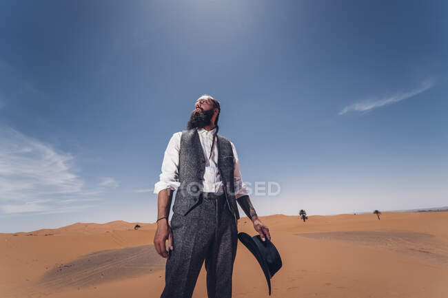 Hombre barbudo disfrazado de vaquero mirando hacia otro lado mientras está de pie en el desierto contra el cielo azul - foto de stock