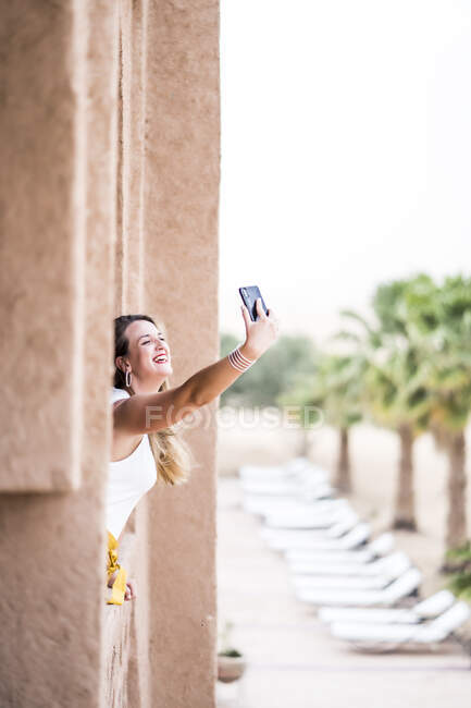 Mujer alegre usando el teléfono móvil para tomar una selfie cerca del paisaje del desierto de pie en el balcón de piedra, Marruecos - foto de stock