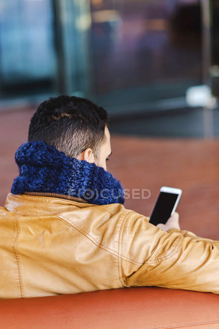 Вид сзади на мужчину в модном наряде, просматривающего смартфон, сидя на удобном кожаном диване — стоковое фото