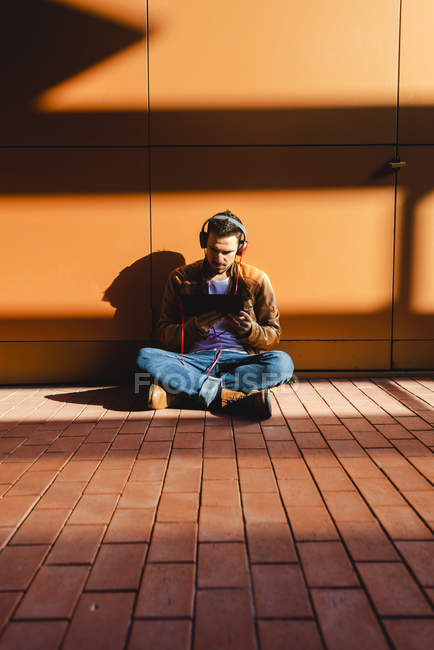 Hombre guapo en auriculares escuchando música y tableta de navegación mientras está sentado en las escaleras fuera del edificio moderno en el día soleado - foto de stock