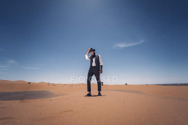 Uomo barbuto in costume da cowboy guardando la fotocamera mentre in piedi nel deserto contro il cielo blu — Foto stock
