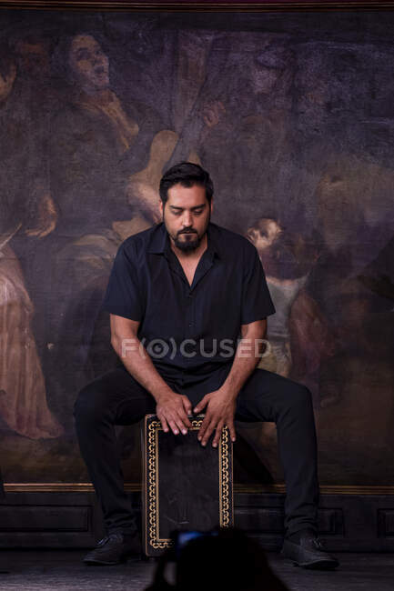 Bearded Hispanic guy playing percussion while sitting against mural during flamenco performance — Stock Photo