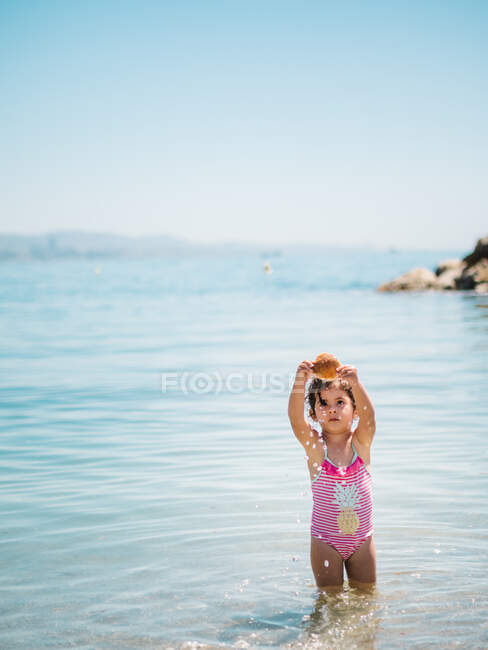 Menina adorável criança em terno de natação em pé em água morna de mar calmo brincando com uma concha — Fotografia de Stock