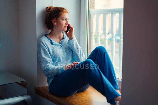 Руда молода жінка в домашньому одязі сидить на підвіконні і розмовляє на мобільному телефоні — стокове фото
