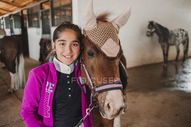 Маленькая девочка обнимается с маленьким пони в милой шляпе на ушах, стоящих внутри конюшни и смотрящих в камеру — стоковое фото
