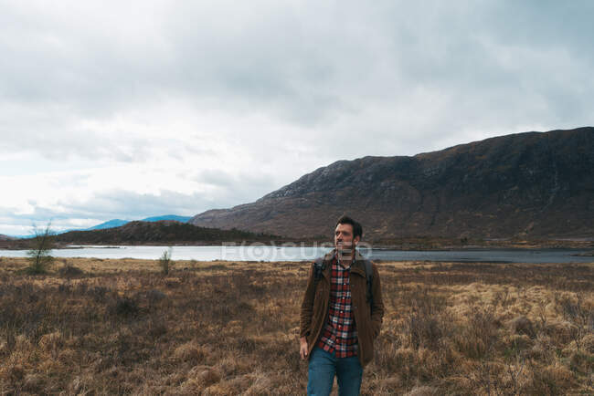 Homme adulte avec sac à dos debout dans une vallée pittoresque et reculée avec des montagnes et un lac regardant loin — Photo de stock