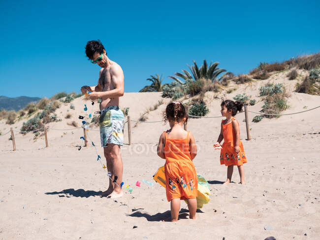 Отец и близнецы в ярко-оранжевых платьях играют с летучим змеем на песчаном побережье в солнечный день — стоковое фото
