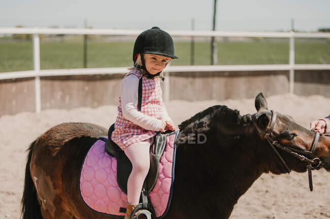 Kleines Mädchen in Kleid und Jockey-Heu sitzt auf dem Pferd und lernt auf der Rennbahn zu reiten — Stockfoto
