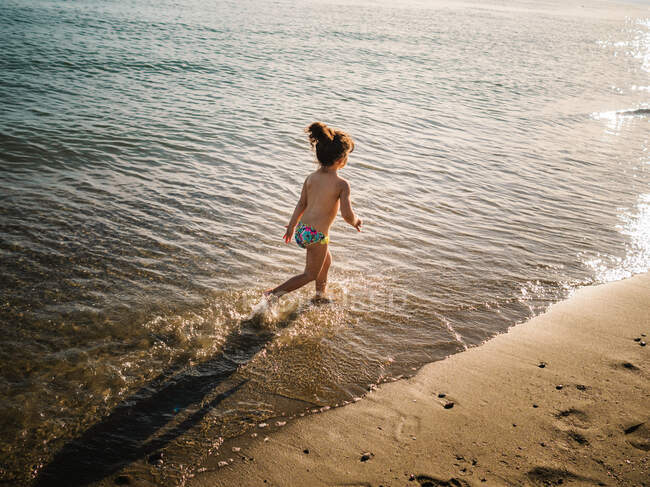 Back view of anonymous cute female kid in swimming slips running on sandy beach against calm sea — Stock Photo
