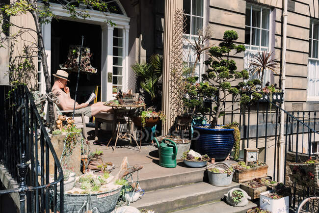 Hombre leyendo el periódico en el porche con plantas creativas y árboles en cajas y utensilios viejos, Escocia - foto de stock