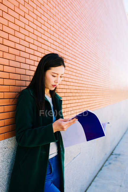 Mujer asiática leyendo libro de texto mientras se apoya en la pared de ladrillo en el campus universitario - foto de stock