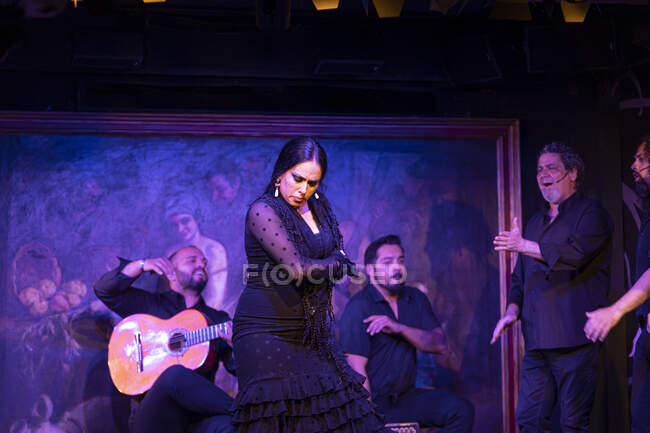 Mujer en traje negro bailando flamenco cerca de músicos masculinos hispanos durante la actuación contra la pintura en el escenario oscuro - foto de stock
