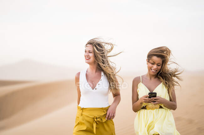 Cheerful stylish two blonde women friends using mobile phone while walking in desert of Morocco — Stock Photo