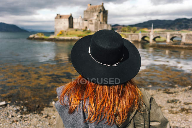 Rückseite einer stilvollen Frau mit Hut, die eine alte Steinburg an der Küste in den Bergen in Schottland betrachtet — Stockfoto