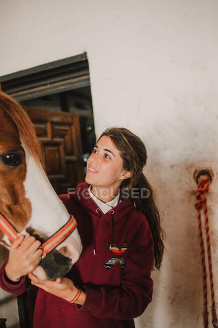 Девочка-подросток обнимается с маленьким пони в милой шляпе на ушах, стоящих внутри конюшни — стоковое фото