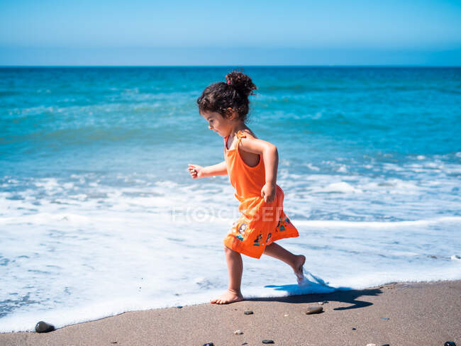 Vue latérale de l'enfant femelle en robe orange courant et jouant sur le bord de mer sablonneux et regardant la mer et le ciel — Photo de stock