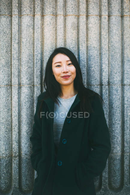 Jovem mulher asiática em casaco elegante sorrindo e olhando para a câmera enquanto se inclina na parede de mármore na rua da cidade — Fotografia de Stock