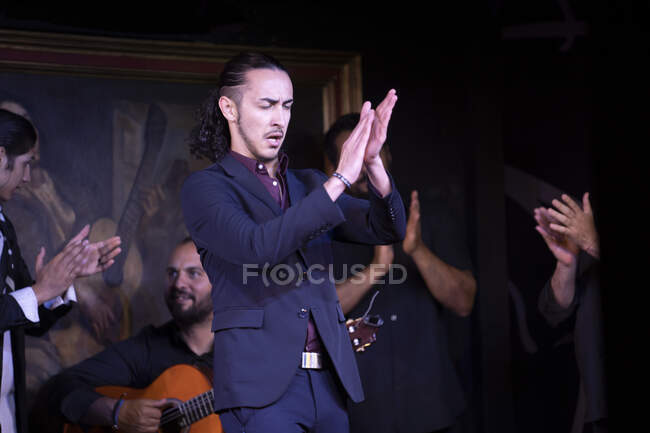 Man in blue costume dancing flamenco near Hispanic male musicians during performance against painting on dark stage — Stock Photo