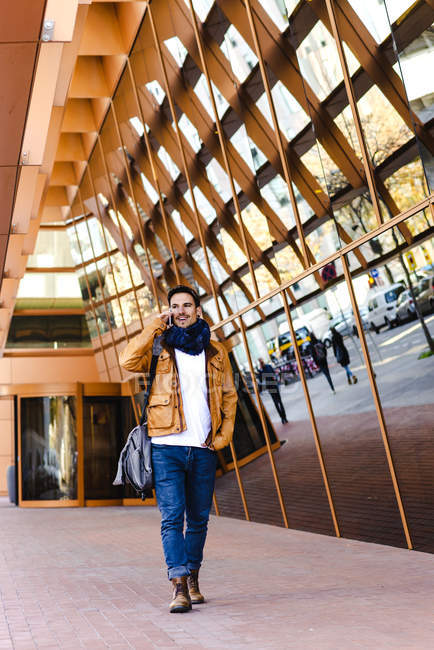 Trendy male in casual outfit talking on mobile phone while walking near glass wall of contemporary building — Stock Photo
