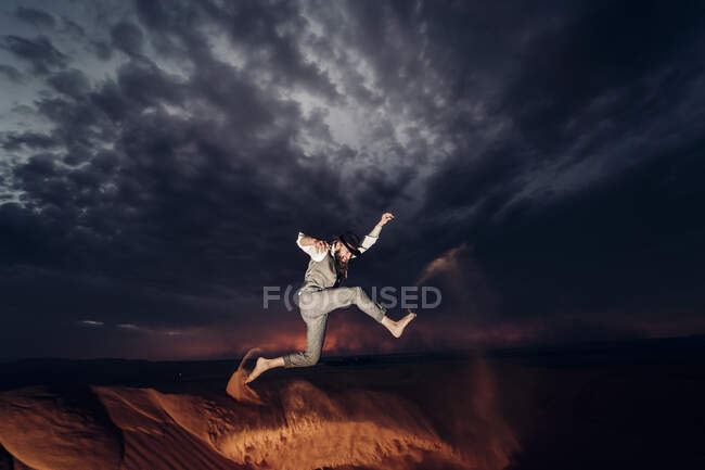 Vista lateral del hombre descalzo en traje de vaquero sonriendo y saltando en el desierto de arena contra el cielo nublado de la noche - foto de stock