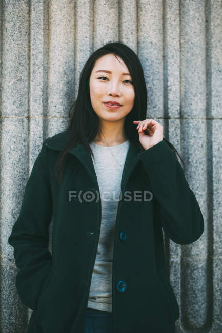 Jovem mulher asiática em casaco elegante sorrindo e olhando para a câmera enquanto se inclina na parede de mármore na rua da cidade — Fotografia de Stock