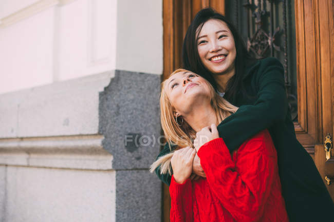 Felice donna asiatica sorridente e in piedi contro la porta ornamentale della costruzione invecchiata e abbracciando amico caucasico — Foto stock