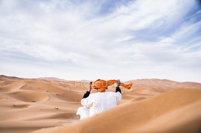 Vista trasera del hombre con traje largo ajustando un turbante colorido sentado en las dunas del desierto arenoso sin fin, Marruecos - foto de stock