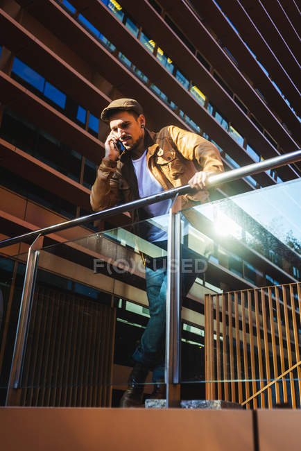 Позитивный человек в стильном наряде разговаривает по мобильному телефону, стоя на современном стеклянном балконе современного здания в солнечный день — стоковое фото