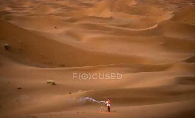 Distant aerial view of stylish brunette setting fire to smoky firework standing in desert of Morocco — Stock Photo