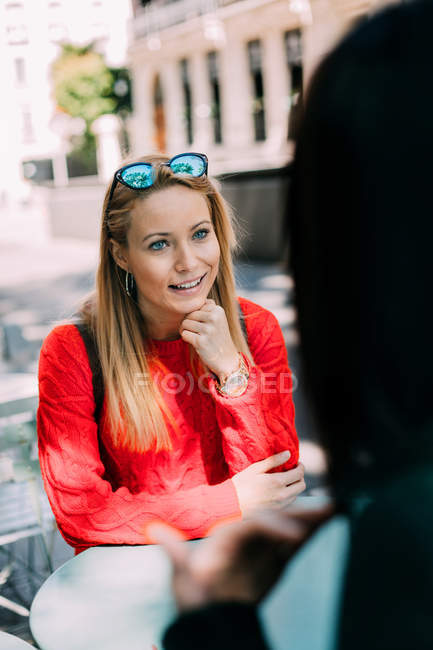 Beautiful young female in knitted red sweater smiling and listening to friend while sitting at table in outdoor cafe together — Stock Photo