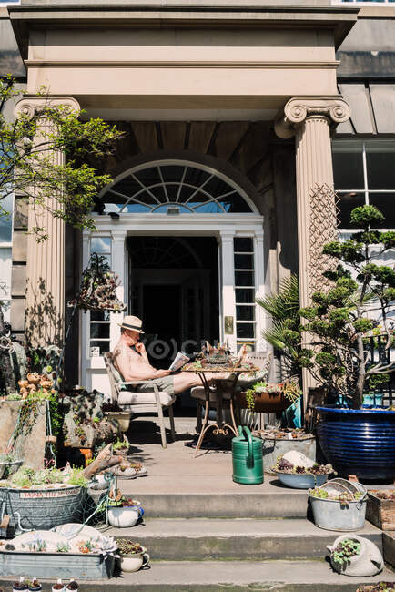 Mann liest Zeitung auf Veranda mit kreativen Pflanzen und Bäumen in Kästen und altem Gerät, Schottland — Stockfoto