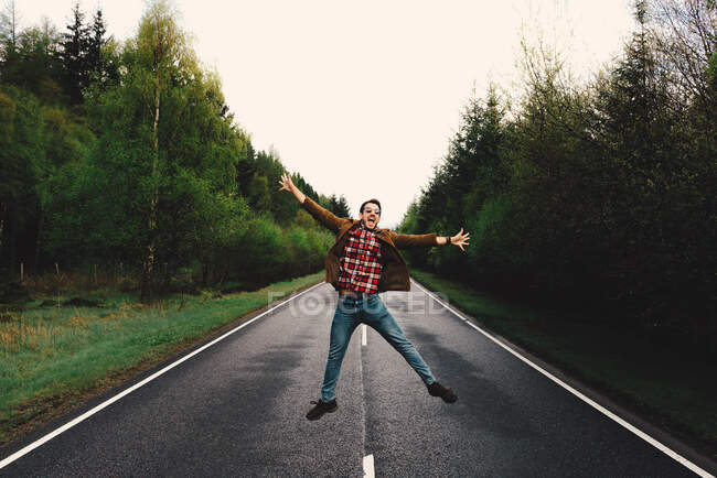 Молодой человек, прыгающий в одиночестве на отдаленной дороге с пышными зелеными деревьями, Шотландия — стоковое фото