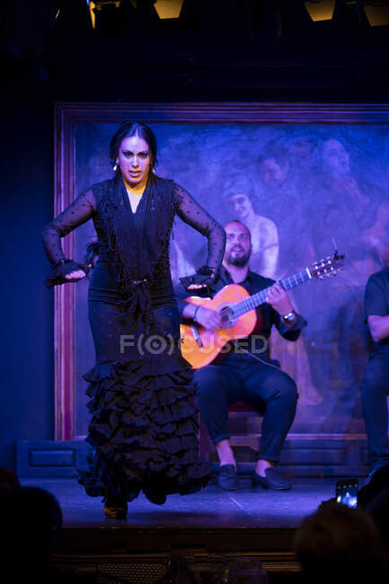 Femme en costume noir dansant le flamenco près des musiciens hispaniques masculins pendant la représentation contre la peinture sur scène sombre — Photo de stock