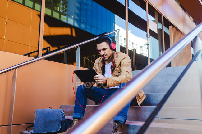Hombre guapo en auriculares escuchando música y tableta de navegación mientras está sentado en las escaleras fuera del edificio moderno - foto de stock