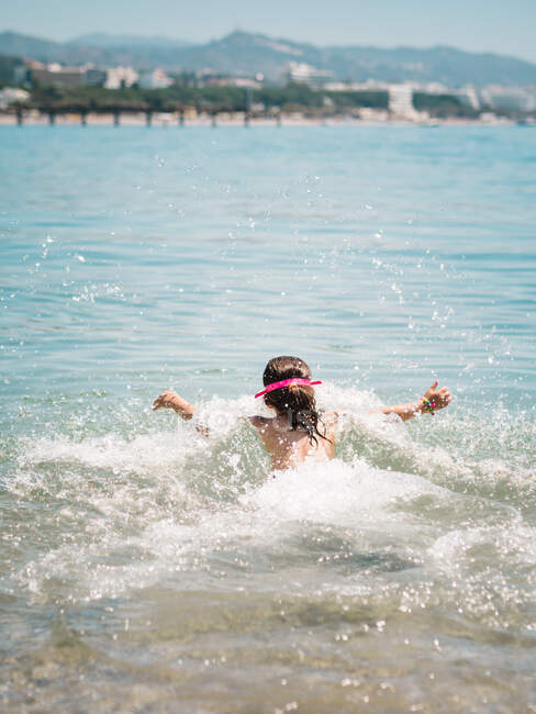 Задний вид анонимной веселой милой девушки, играющей в воде моря на фоне спокойного побережья — стоковое фото