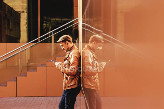 Позитивный человек в стильном наряде с помощью мобильного телефона, опираясь на стену современного здания в солнечный день — стоковое фото